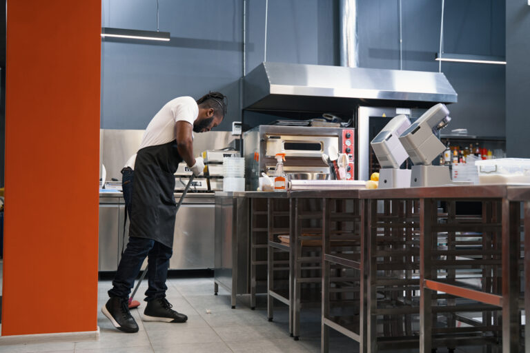 African man in uniform standing in the kitchen, holding mop and cleaning up after work, looking down