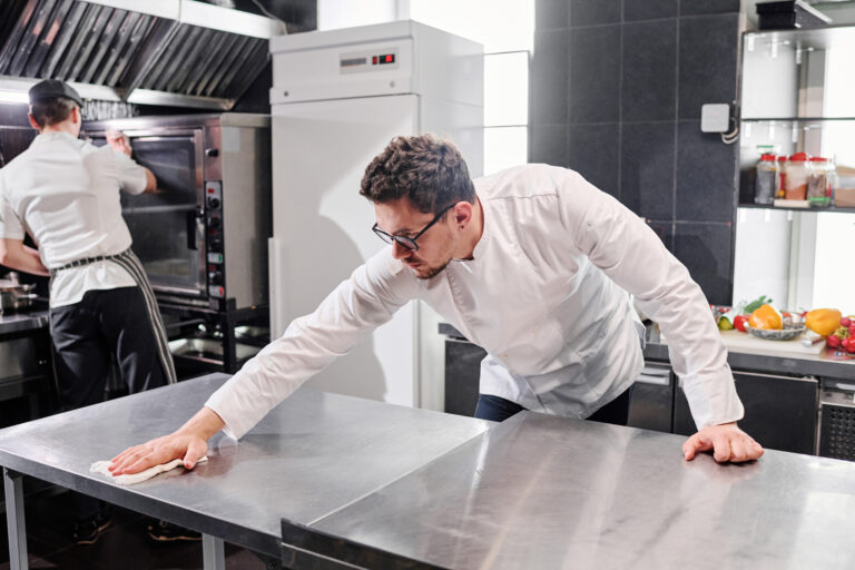 Chef in uniform wiping kitchen table after cooking with his colleague cleaning the oven after work in kitchen