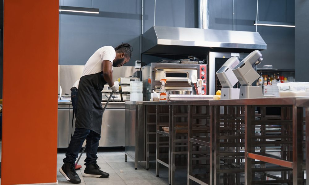 African man in uniform standing in the kitchen, holding mop and cleaning up after work, looking down