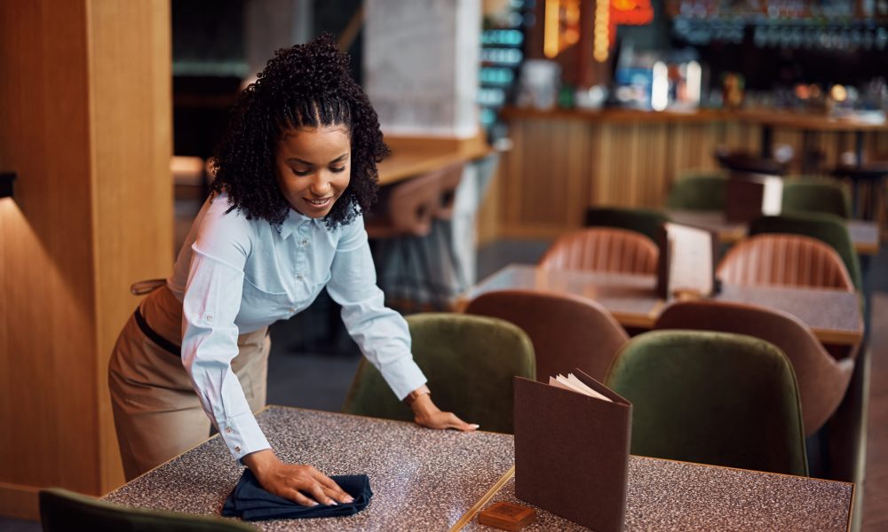 Young African American waitress cleaning tables while working in restaurant.