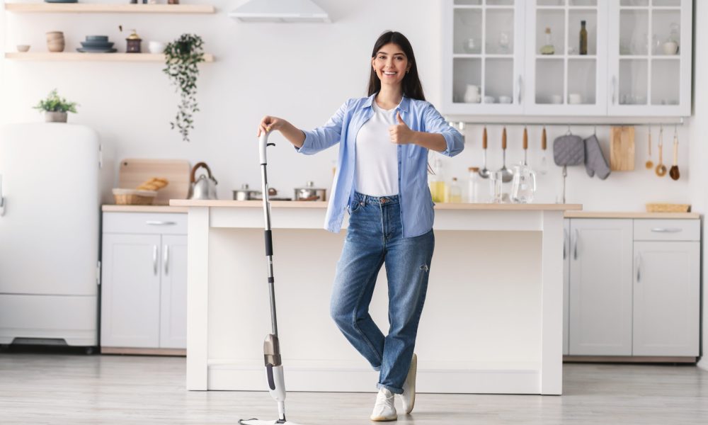 Smiling woman showing thumb up sign gesture after washing hardwood laminate flooring, holding and leaning on water spray mop pad, standing at kitchen posing at camera, full body length