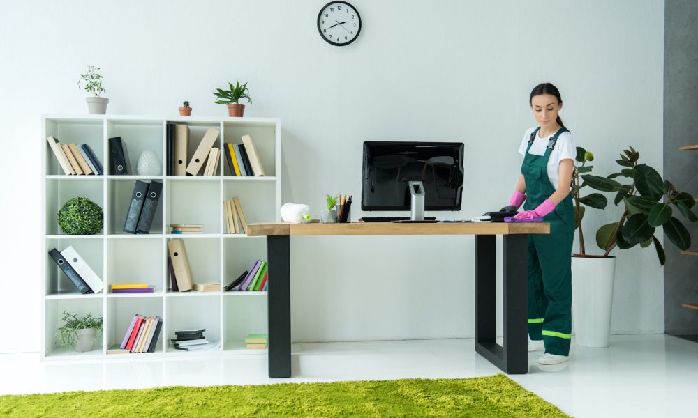 young cleaning company worker in rubber gloves and uniform cleaning desk in modern office